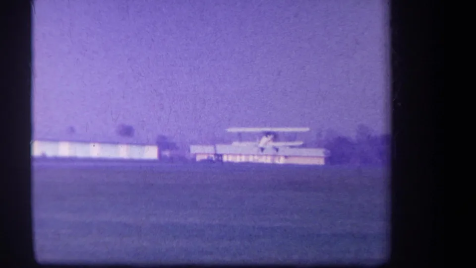 1959 Volmer Yucaipa frame with a small biplane gaining speed across a rural airfield, backed by low buildings and a distant tree line.