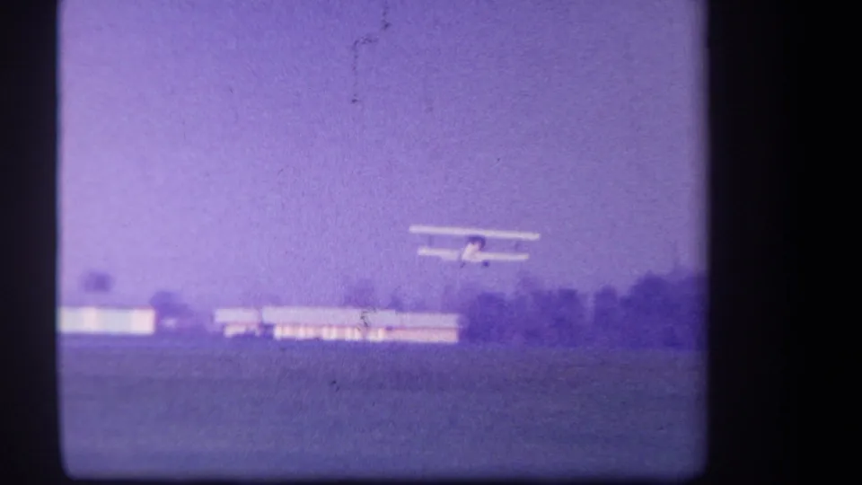 1959 Volmer Yucaipa 8mm frame showing a small biplane just airborne above a grass airfield, with buildings and trees below the horizon.