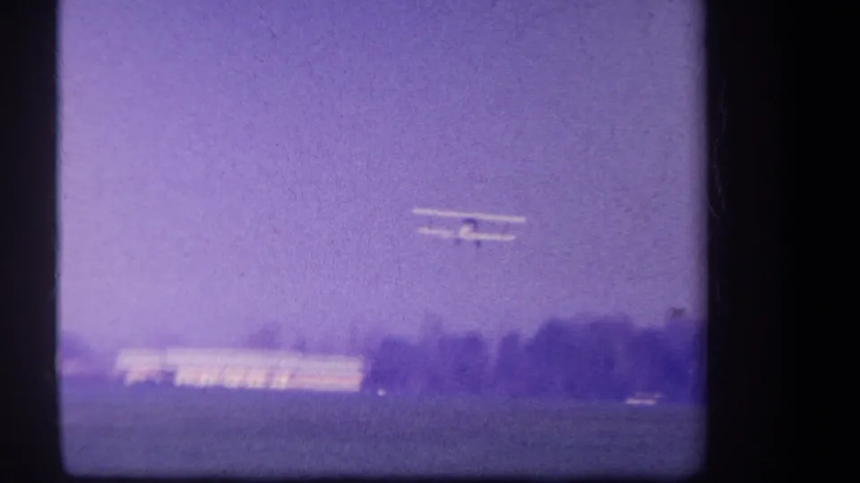 1959 Volmer Yucaipa frame showing the small biplane airborne above the distant field, with low buildings, trees, and purple-tinted sky visible.