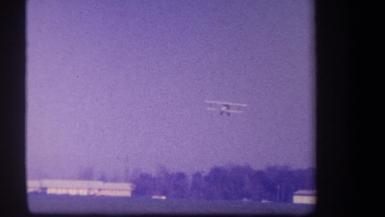 The biplane remains high in frame over a rural California field, with trees and light-colored buildings along the horizon.