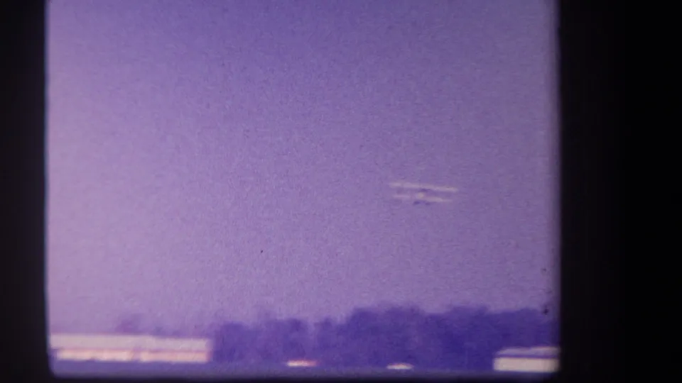 A small biplane is faintly visible in a grainy purple sky above the distant rural horizon in the 1959 8mm footage.
