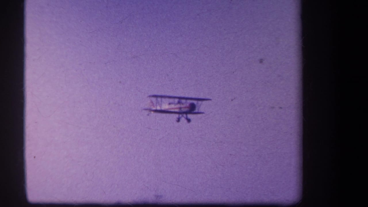 Small biplane flying against a hazy purple sky with dark film borders.