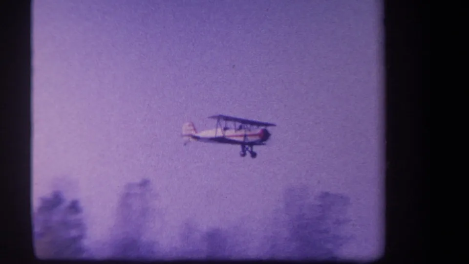 White and red biplane passes above blurred trees in 1959 rural Yucaipa home-movie footage.