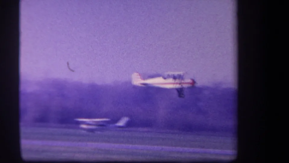 Low pass by a white and red biplane near a rural field, with a distant aircraft-like shape below in the 1959 footage.