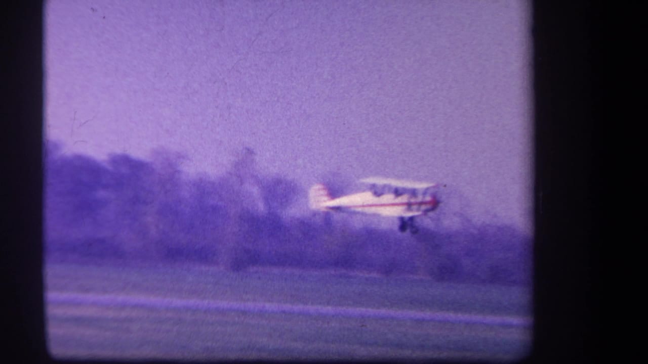 1959 Yucaipa-area home-movie frame showing a small white prop plane low over a grass runway, with trees and a hazy sky behind it.