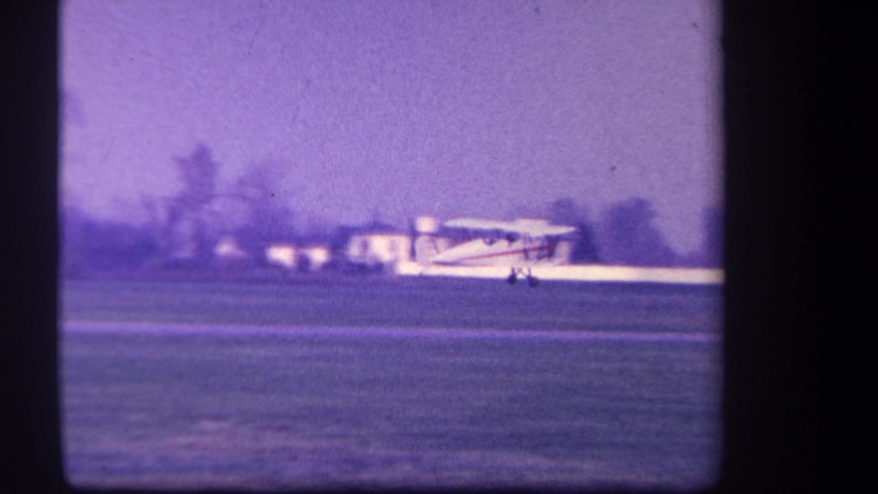 Small white plane near touchdown on a grassy airstrip with low buildings beyond.