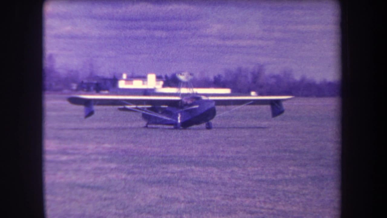Small propeller plane parked on a grass airfield with distant buildings and trees.