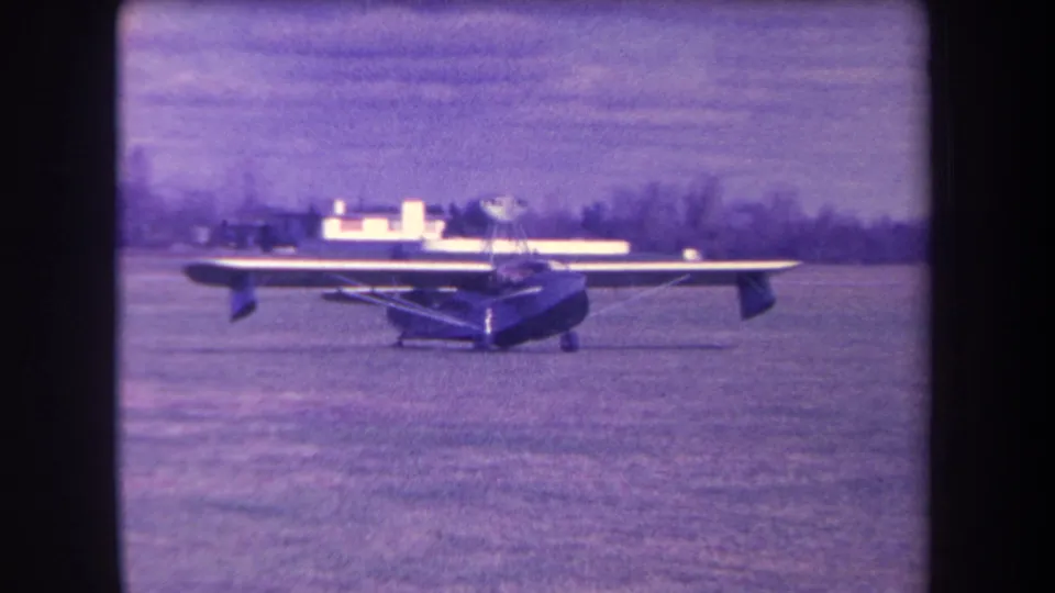 1963 8mm frame from Yucaipa area showing a small high-wing propeller plane stationary on a grassy airfield.