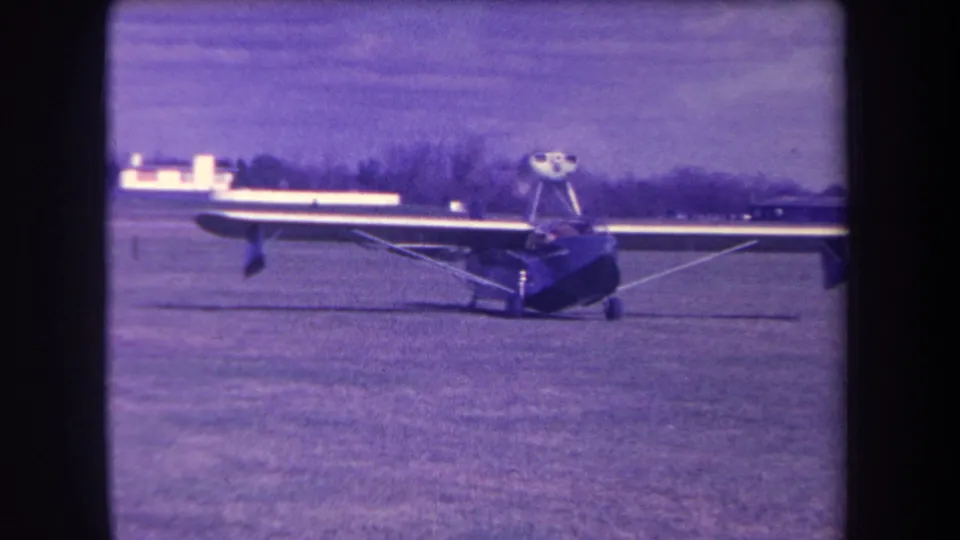 1963 8mm frame with the parked high-wing light plane filling more of the view on a grass airfield.