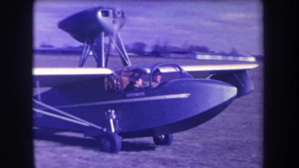 1963 Yucaipa home-movie frame with a side view of the small amphibious aircraft and two people seated in the cockpit.