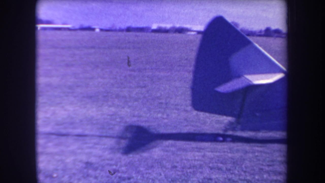 A small aircraft's tail and rotor area appear against the grass airfield, with distant low structures beyond the field.