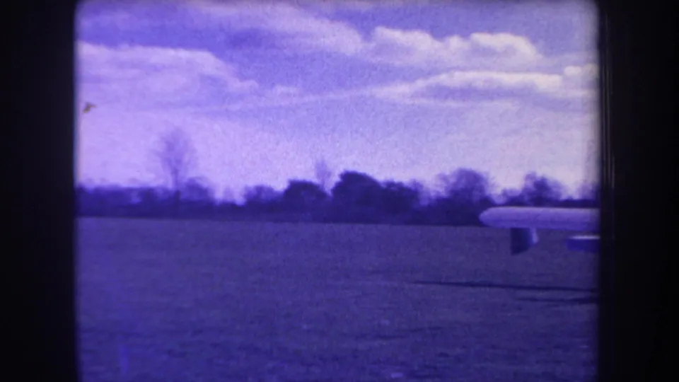 The aircraft moves partly out of frame, leaving a wing edge and landing gear visible against the 1963 grass airfield and cloudy sky.