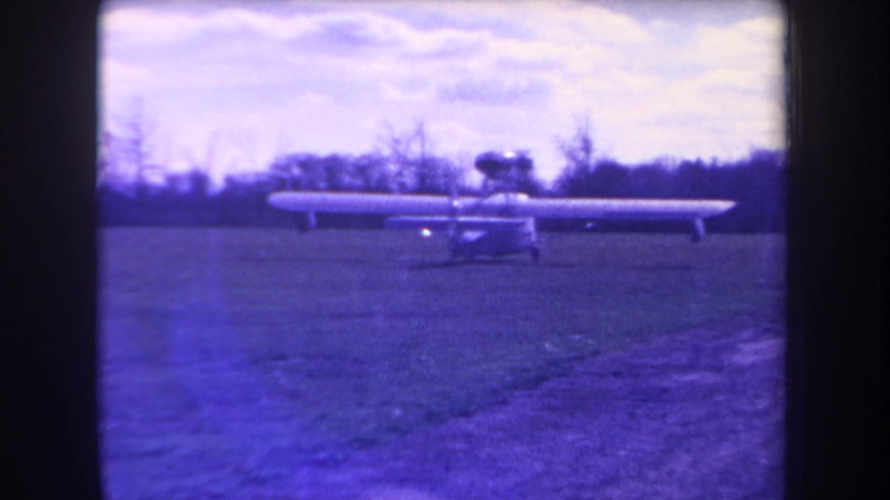Side view of a small long-wing aircraft on a grassy Yucaipa field, with purple fading from aged 8mm film.