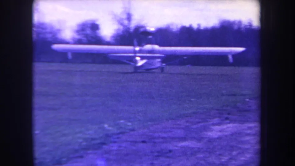 1963 Yucaipa-area home-movie frame showing a small propeller plane on a grass field with a dark tree line and pale sky behind it.