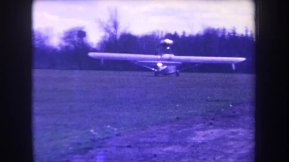 1963 archival home-movie still of a light single-engine airplane on grass, with wooded background and visible 8mm grain.