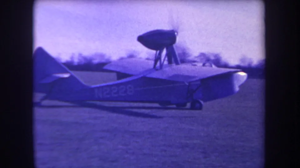 1963 Yucaipa frame showing the amphibious aircraft continuing across the field, with the raised propeller pod silhouetted against a soft tree line.