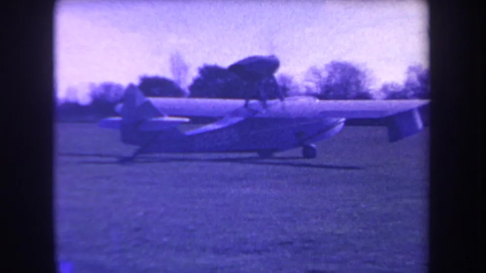 Archival 8mm frame of the small aircraft on a grassy California field, with propeller blur and soft tree silhouettes behind it.