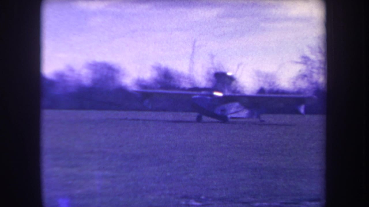 Small amphibious plane on a grassy field with dark film borders and trees in the background.