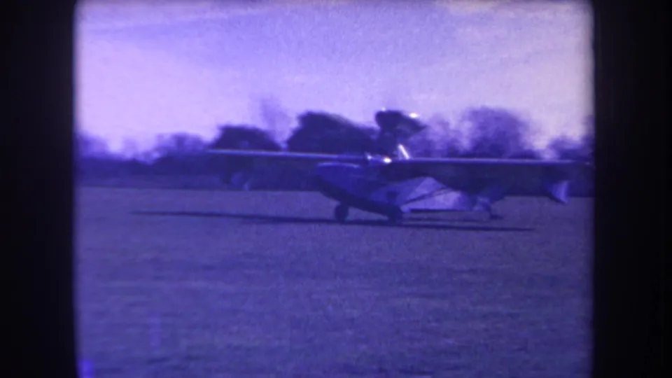 Side view of a small amphibious aircraft rolling across a field in 1963 home-movie footage, with trees beyond the field.