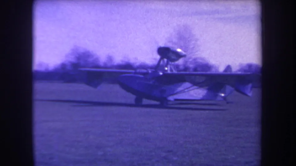 Closer 8mm view of the amphibious aircraft on the field, showing its raised engine pod and boat-like fuselage.
