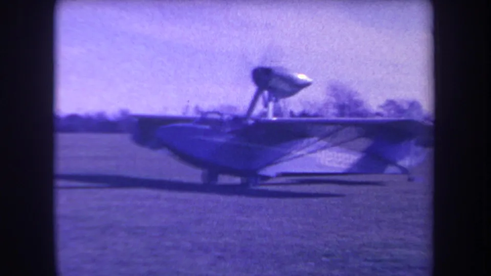 The small amphibious aircraft fills more of the frame as it moves across the grass, with purple-tinted 8mm film color.