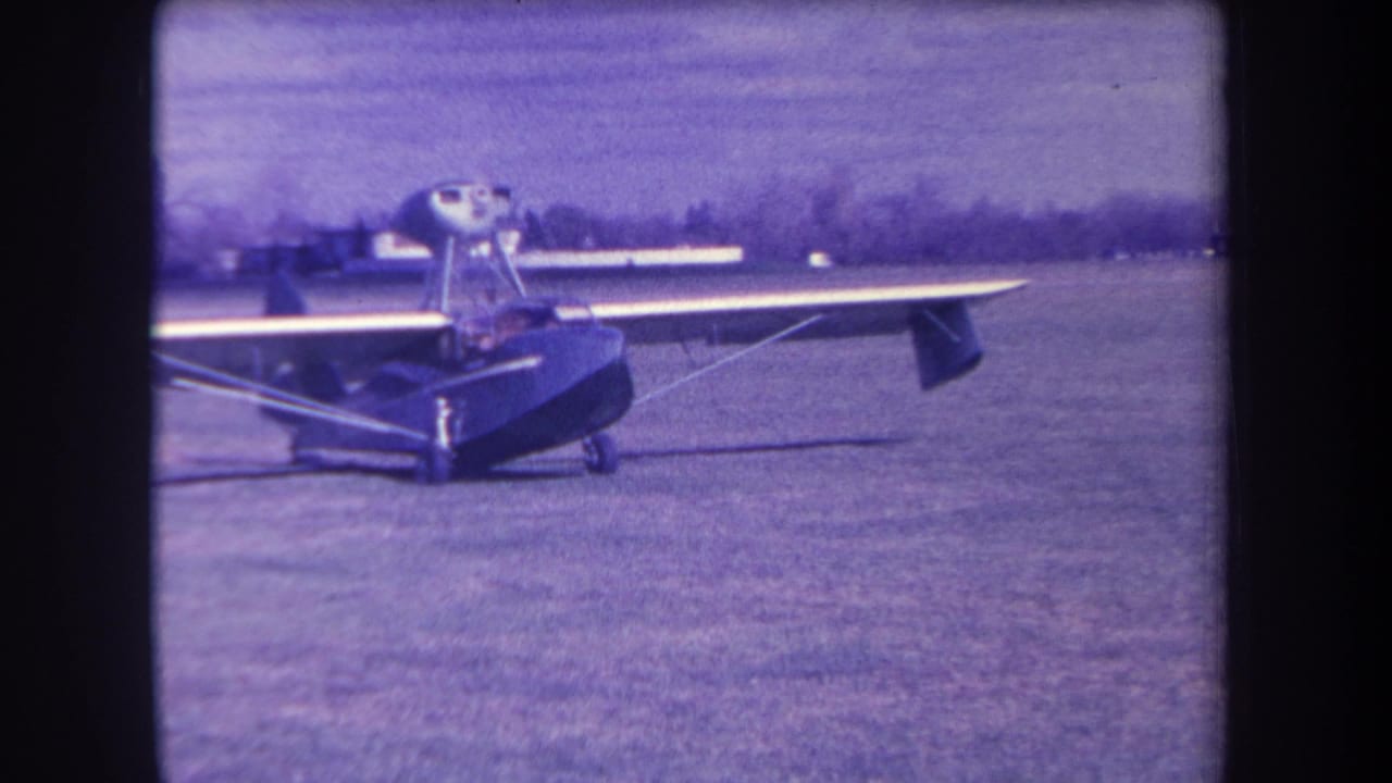 1963 8mm frame from Yucaipa, California, showing a dark high-wing amphibious aircraft stationary on a grassy airfield.