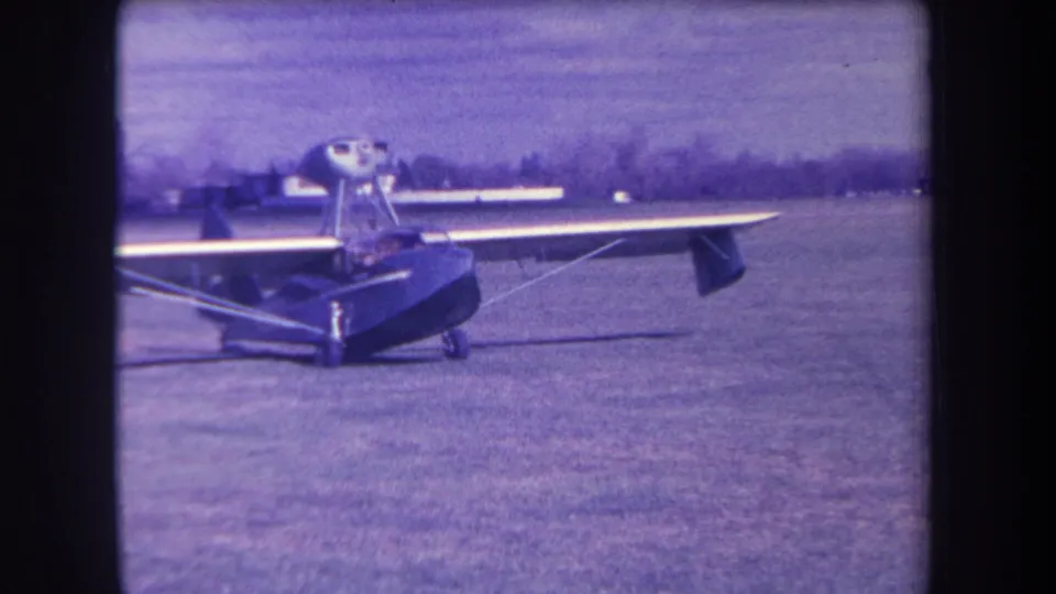 1963 8mm frame from Yucaipa, California, showing a dark high-wing amphibious aircraft stationary on a grassy airfield.