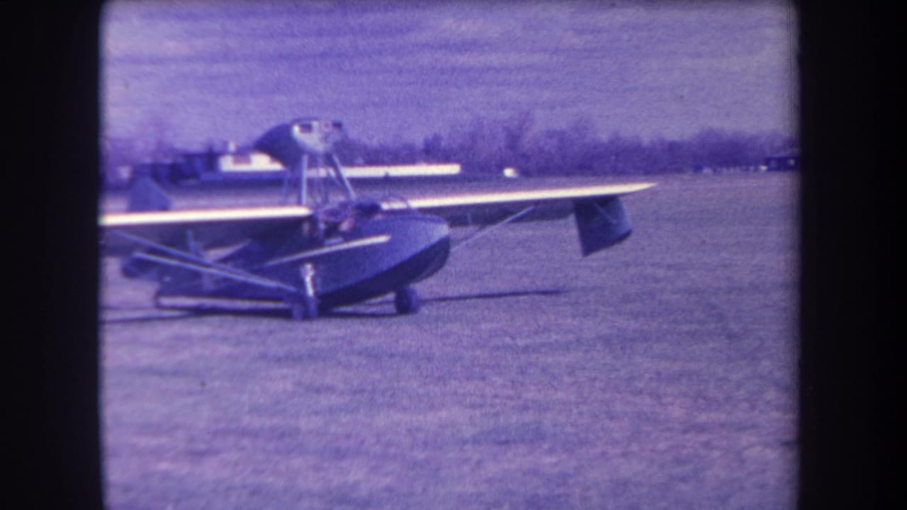 High-wing amphibious plane seen from the front side on a grassy airfield.