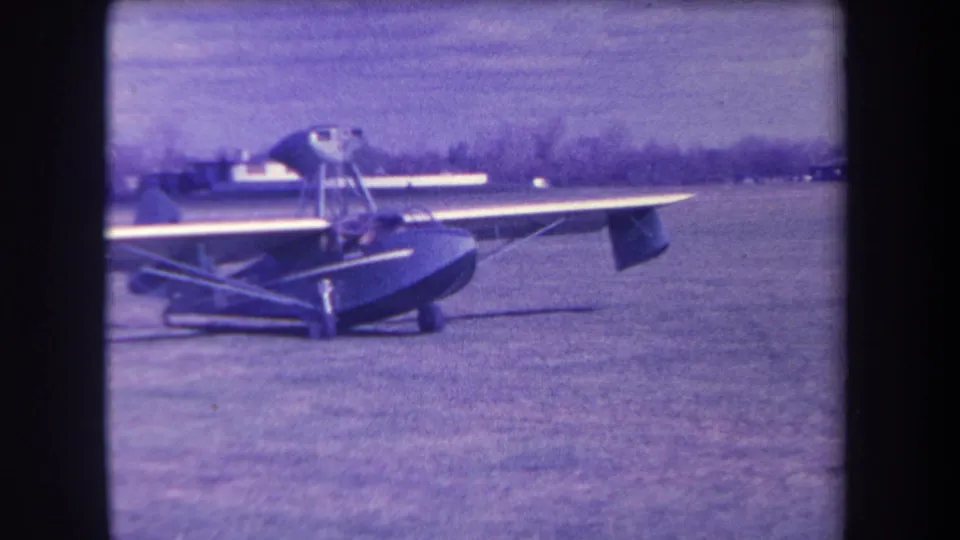 The small amphibious aircraft remains parked on the grass in this 1963 Yucaipa home-movie frame, with a person faintly visible in the cockpit area.