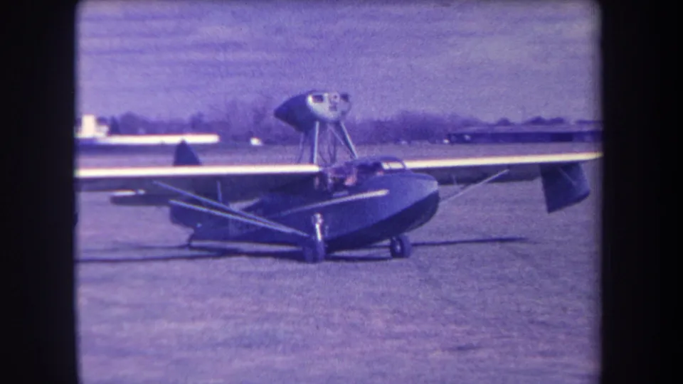 Final 1963 Yucaipa frame of the dark amphibious aircraft parked on the grass airfield, with trees and low buildings beyond.