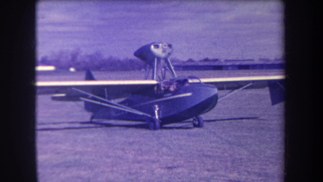 Small amphibious aircraft parked on a grass field with a pilot in the cockpit.
