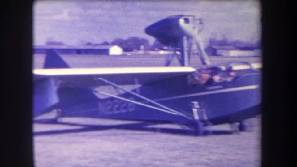 Close 1963 home-movie frame of the amphibious aircraft side, with cockpit, pilot, wing struts, and registration markings visible.