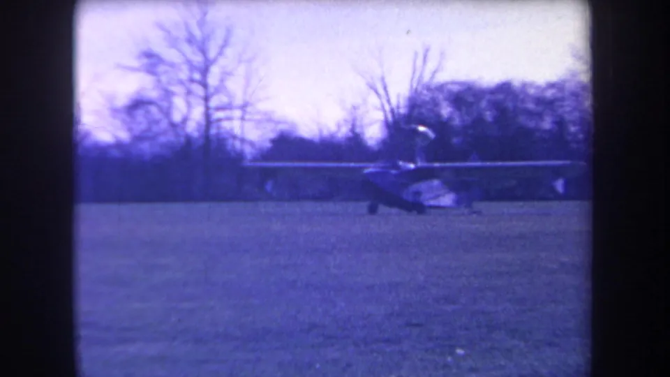 Side view of a small single-engine plane on a rural grassy field, captured in blue-shifted 1963 8mm footage.