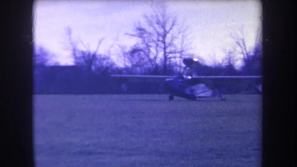 Final blue-tinted frame showing the small aircraft on grass at Volmer Yucaipa, with bare trees and dark scan borders visible.