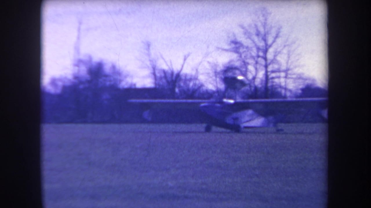 Small single-engine plane moving across a grass field with bare trees behind it.