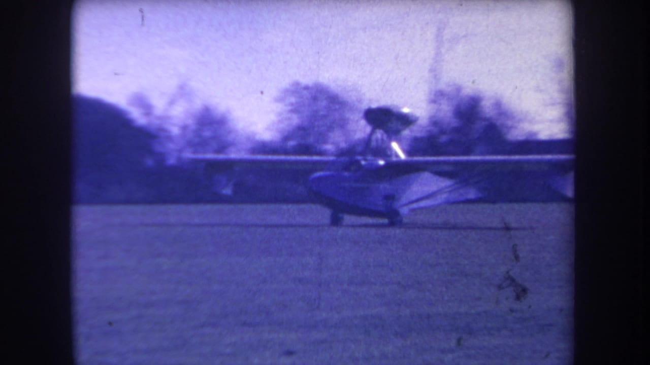 The plane continues across the Volmer Yucaipa grass field, seen through soft blue-purple 8mm film color.