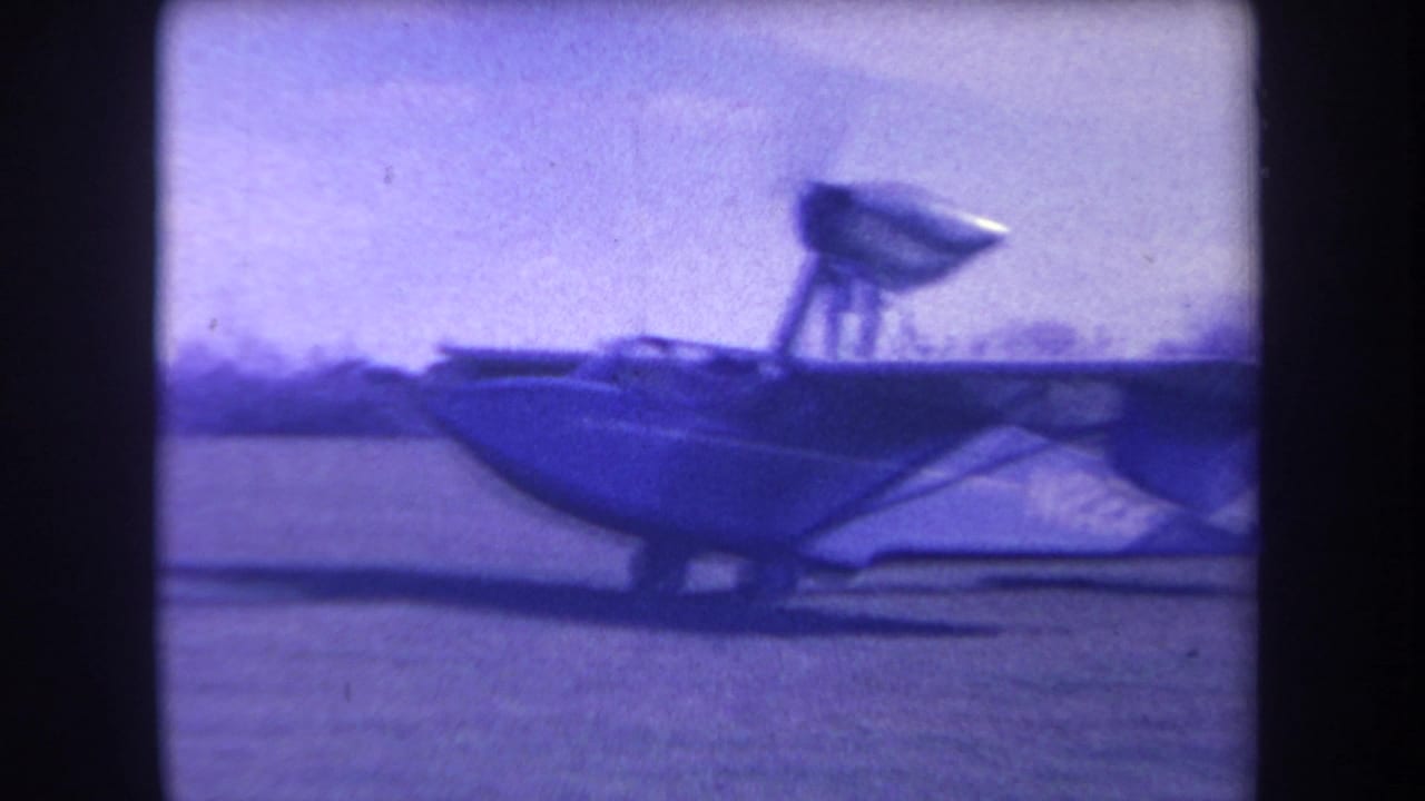 Blue amphibious aircraft seen side-on on a grassy airfield.