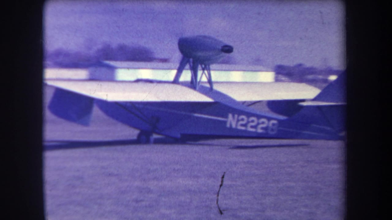 Small airplane marked N2226 on a grass airfield, seen through an aged 8mm frame.