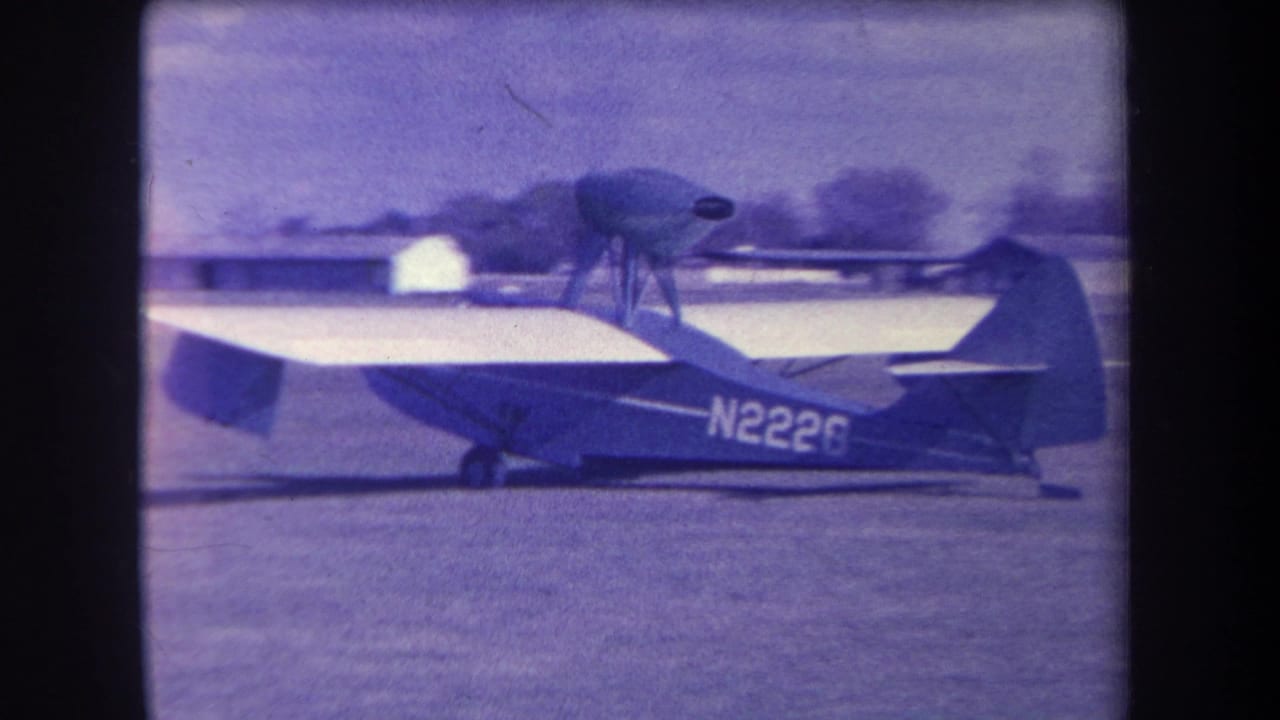 1963 archival home-movie frame of the N2226 aircraft taxiing on a grass airfield near Yucaipa, with distant buildings and soft film grain.