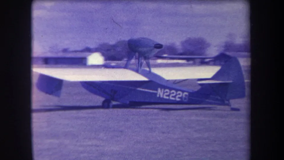 1963 archival home-movie frame of the N2226 aircraft taxiing on a grass airfield near Yucaipa, with distant buildings and soft film grain.