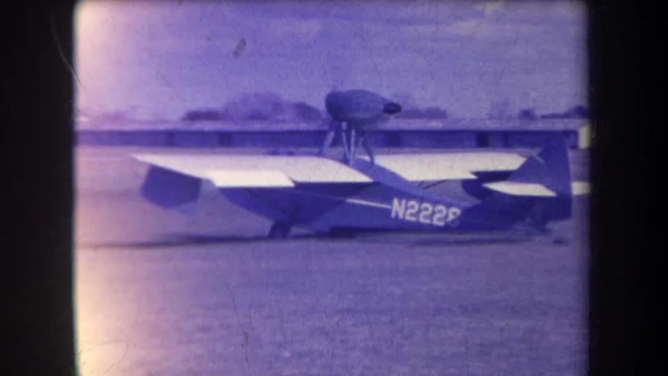 1963 Yucaipa-area 8mm frame showing the N2226 light aircraft in a turn on grass, with blurred airfield structures in the background.