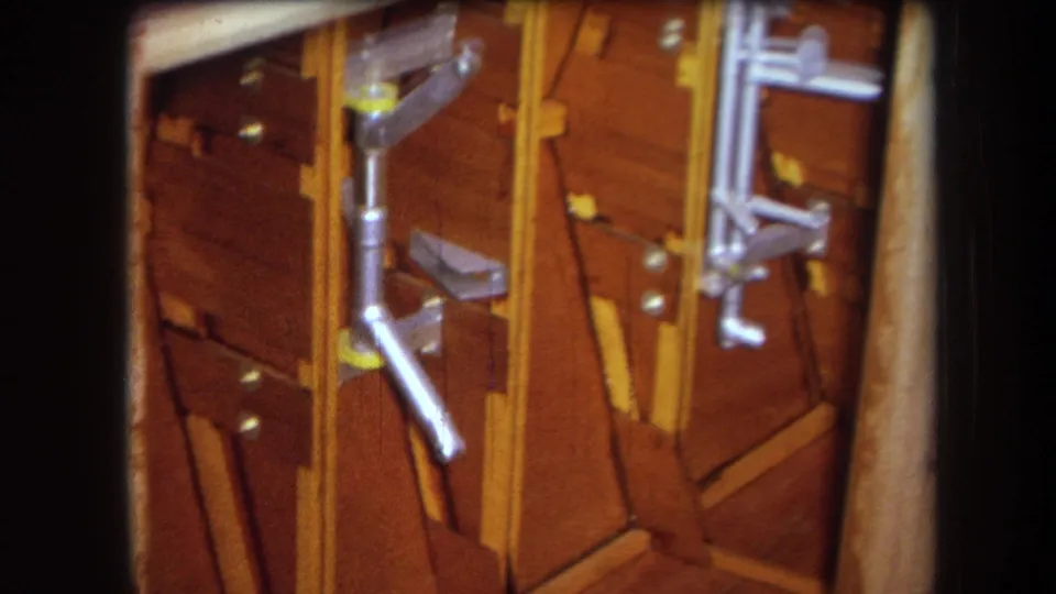 1964 8mm frame with a tilted handheld view of varnished wood braces, silver rods, brackets, and small hardware inside an apparatus.