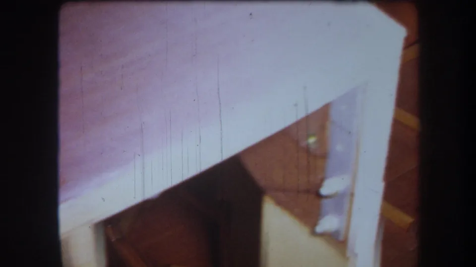 1964 home-movie frame showing a white-painted counter or table edge, wood floor, and part of a wooden cabinet-like structure below.