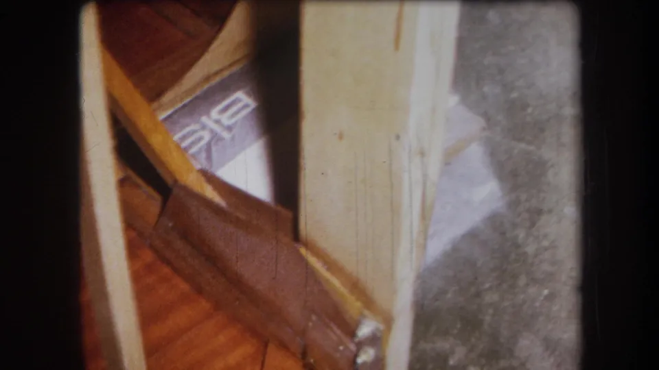 1964 8mm frame from Yucaipa showing a lower cabinet or table corner with wood supports, pale paneling, and gray floor surface.