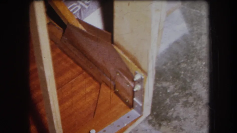 1964 Yucaipa frame showing a wooden cabinet or table corner with pale side panel, dark rail, visible bracket hardware, and gray floor.