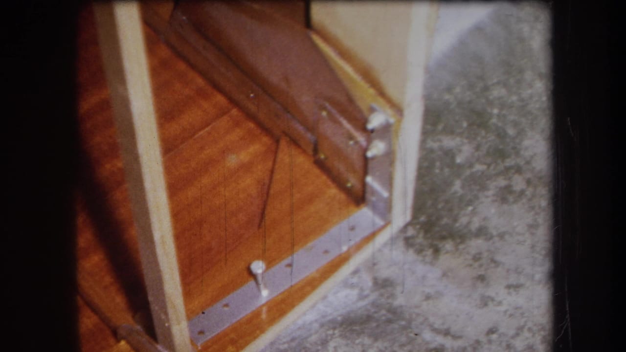 The frame shows an angled close-up of wood damage, a metal strip with screws, pale vertical trim, and rough gray flooring.