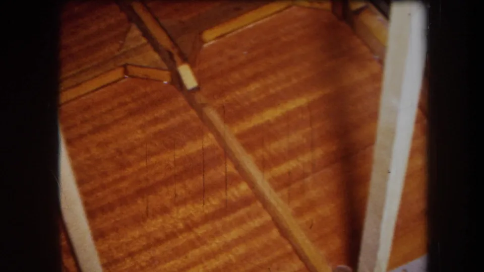 Close 8mm view of a warm-toned wooden panel or table surface with a loose narrow piece and pale structural edge nearby.
