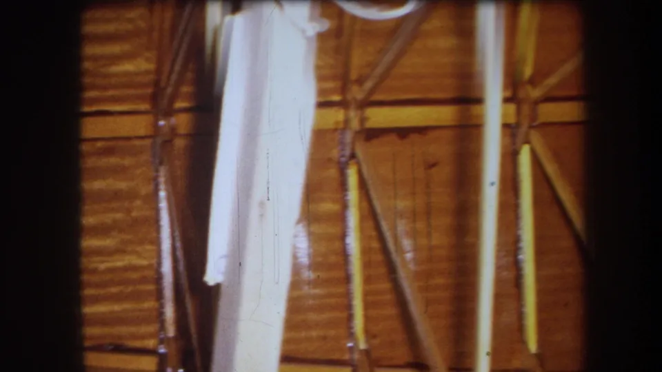1964 8mm view of a wooden crawl space or attic area, including exposed supports, amber wall panels, and a pale hanging fabric-like form at left.