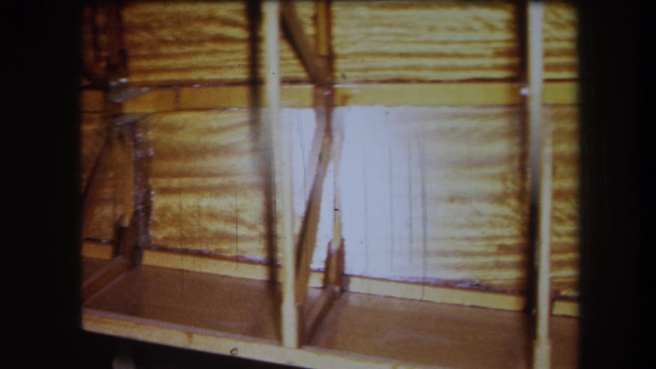 Unfinished wooden boat side with vertical supports and planking in a workshop.