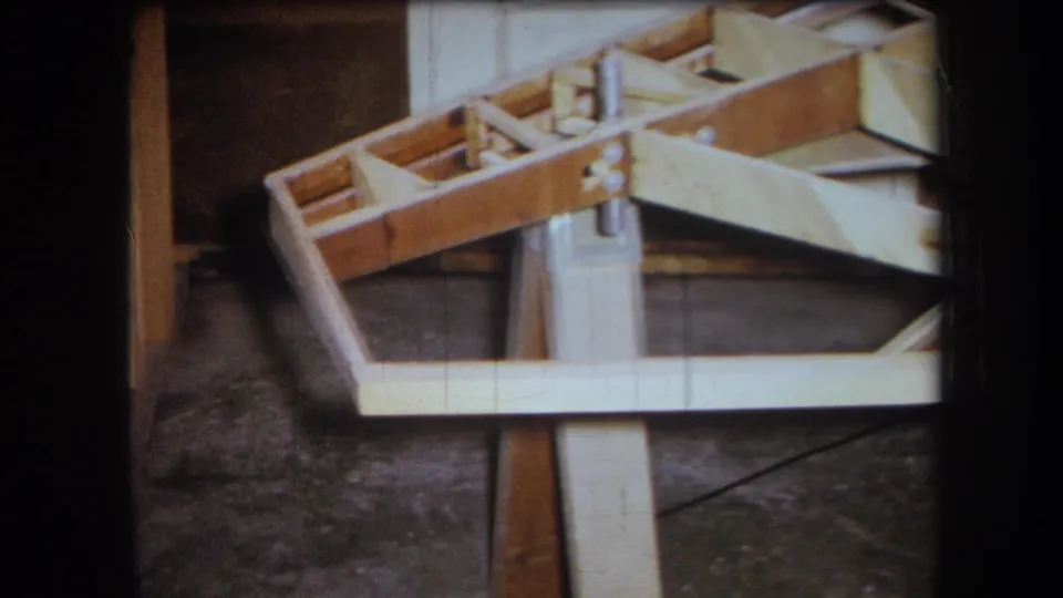 Close 1964 Yucaipa 8mm view of an unfinished wooden boat frame, showing ribs, supports, and workshop floor beneath.
